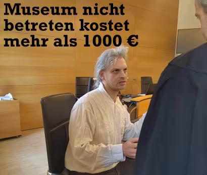Christian Bläul sitzt im Landgericht. Über ihm ist Schrift zu sehen „Museum nicht betreten kostet mehr als 1000 €“